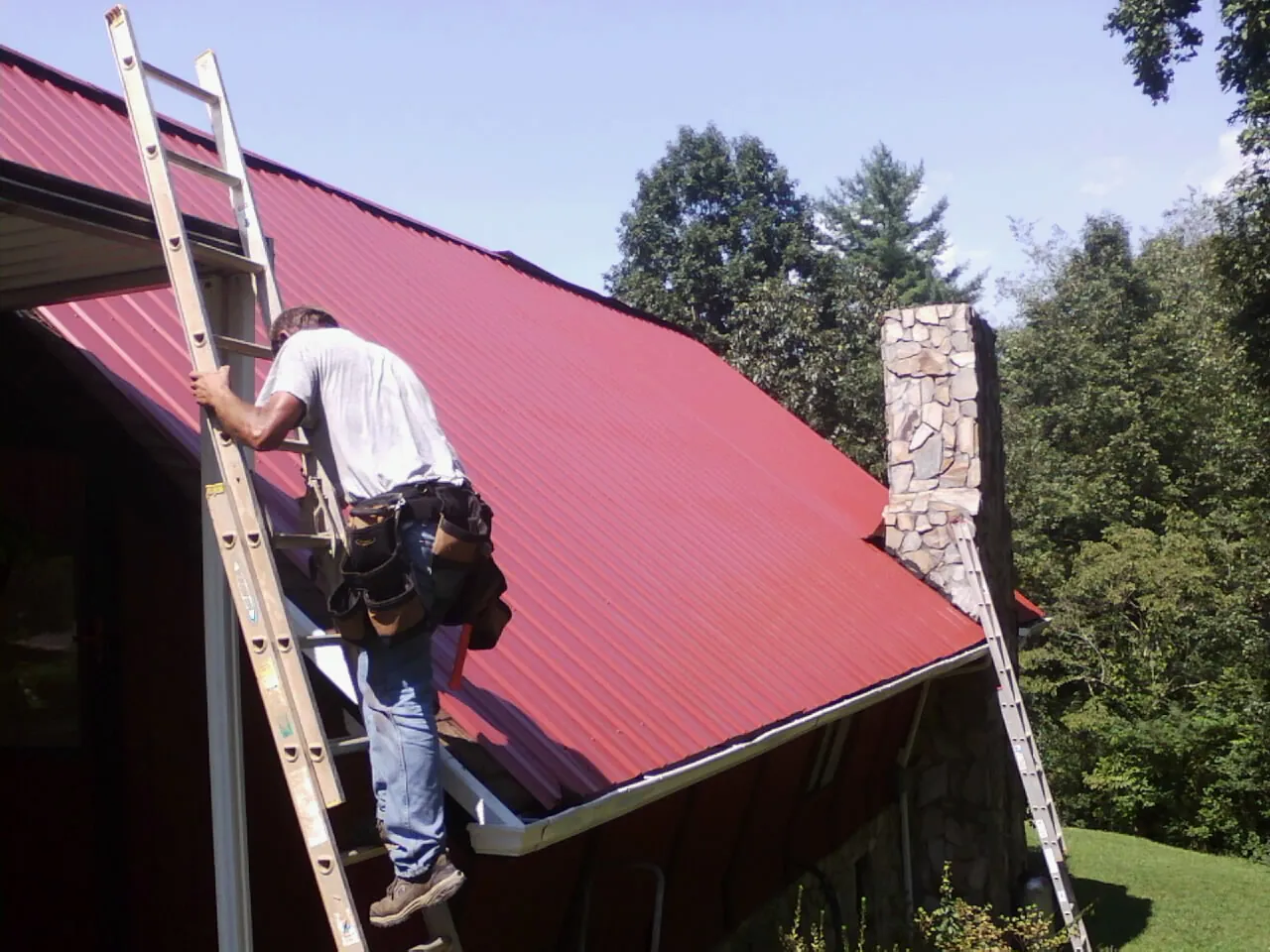 Roofer on a ladder beside a red metal roof and stone chimney — Soffit & Fascia Repair by Roofing Brothers in Attleboro